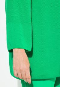 Grön tröja med texturerad stickning. Löst snitt med vida ärmar. Hand synlig som vilar på kanten. Mjuk yta, livfull färg.