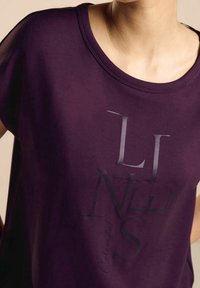 Camisa corta de manga en color púrpura con escote redondo que presenta un estampado de texto negro en capas. Tejido suave con un ligero brillo.