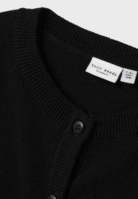 Schwarzer Cardigan mit einem gerippten Ausschnitt und Knopfleiste. Verfügt über eine glatte Strickstruktur und ein Etikett mit der Aufschrift "basic needs by name it."