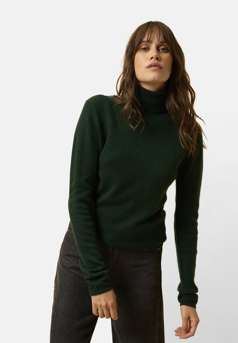 Maglione a collo alto verde scuro realizzato in tessuto morbido e di tipo testurizzato, con maniche attillate, indossato sopra pantaloni scuri. Design semplice senza motivi.