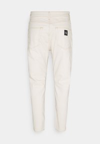 Béžové džínové kalhoty se rovnými nohavicemi, dvěma zadními kapsami a černou nášivkou s logem. Tkanina má hladkou strukturu a viditelné švy.