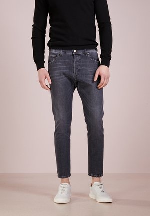 Man wearing faded black slim-fit jeans, white sneakers, and a black long-sleeve sweater standing against a plain background.