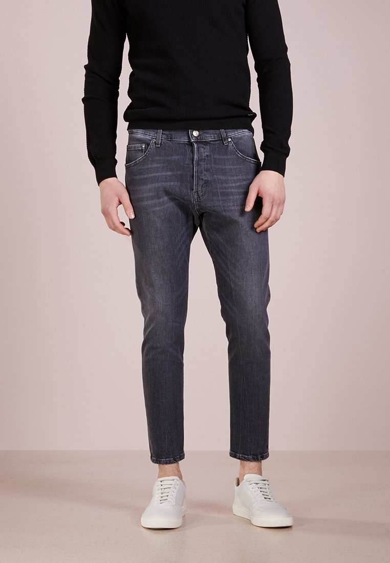 Homme portant un jean noir slim délavé, des baskets blanches et un pull noir à manches longues, debout devant un fond uni.