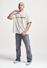 Light beige cotton t-shirt with "2Y STUDIOS" printed in black. Worn with gray denim jeans and blue-and-white sneakers.