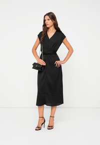 Black wrap dress with short sleeves, knee-length, smooth fabric, and a tie at the waist. Paired with black strappy heels and a small handbag.