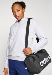 Mala de desporto preta com o logótipo "adidas" branco, feita de material resistente; possui fecho de zíper e alça ajustável. Modelo a usar um moletom cinza.