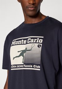 Ναυτικό μπλε βαμβακερό T-shirt με γραφικό σχέδιο ενός τενίστα σε κρεμ απόχρωση, με κείμενα «Monte Carlo» και «JÖRN BORG Tennis Club» συνοδευόμενα από ορ�ιζόντιες γραμμές.