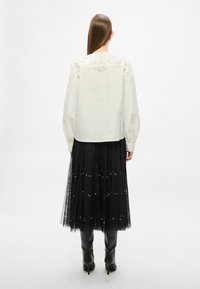 Biela blúzka s detailnou výšivkou na ramenách, v kombinácii s čiernou čipkovanou sukňou s perlami a vysokými čiernymi čižmami.