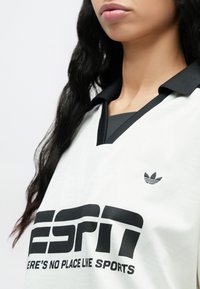 Bela športna majica z oznako ESPN s črnimi poudarki, na prsih ima velik črn logotip in majhen Adidasov znak, z klasičnim polo ovratnikom.