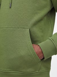 Persoon die een groene hoodie draagt met één hand in de voorste kangoeroezak, met zicht op geribde boord en trekkoorddetail.