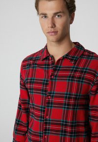 Rot-schwarz kariertes Hemd mit Knopfdesign, Kragen und türkisfarbenen Akzenten im Muster. Weicher Stoff.