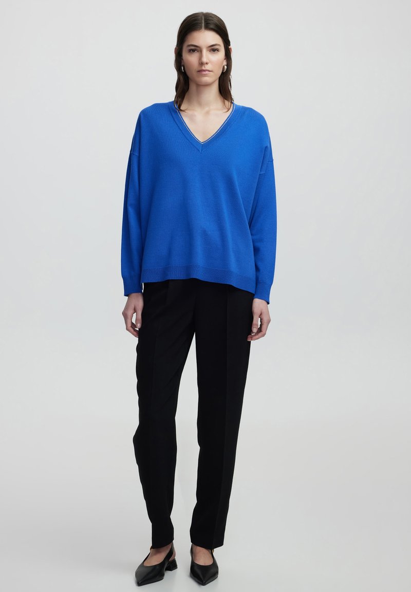 Blauer, übergroßer Pullover mit V-Ausschnitt, strukturiertem Strick, kombiniert mit schwarzen Tailor-Hosen und spitzen schwarzen Ballerinas. Schlichte, klare Linien.