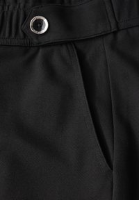 Mustan kankaan lähikuva, jossa näkyy nappikiinnityksellä varustettu vyötärö ja sivutasku, joissa on yksityiskohtainen ompelutekniikka housuissa.