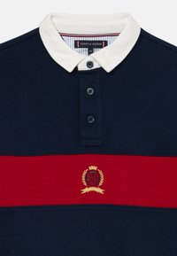 Polo de color marino con cuello blanco, que presenta una franja horizontal roja y un escudo bordado en oro con un león y una corona. Con tapeta de cuatro botones.