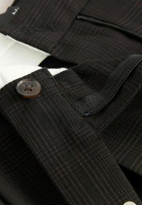 Tessuto nero a motivo con dettagli in marrone scuro, dotato di chiusura a bottone e zip. La texture appare liscia con un delicato dettaglio a griglia.