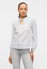 Νεαρή γυναίκα που φοράει λευκό μπλουζάκι με μπλε λουλουδάτο σχέδιο και γιακά με βολάν, σε συνδυασμό με ανοιχτό γκρι τζιν, στέκεται μπροστά σε λευκό φόντο.