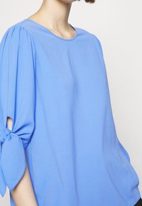 Top azzurro chiaro con maniche corte e nodi legati ai gomiti. Tessuto liscio, collo rotondo e vestibilità comoda. Design semplice e arioso.