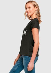 Schwarzes Baumwoll-T-Shirt mit kurzen Ärmeln, das ein bedrucktes Design auf der Vorderseite zeigt. Über blauen Jeans getragen, mit einem lässigen Schnitt und Seitenansicht.