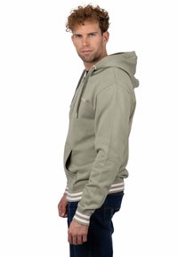 Sudadera con capucha de color verde claro, realizada en tejido suave. Cuenta con un bolsillo tipo canguro y puños y bajo de canalé con rayas. Mostrada puesta por un modelo.