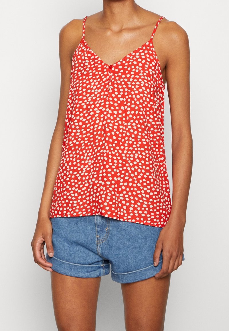 Haut rouge sans manches à pois blancs, encolure en V et détail de boutons à l'avant. Tissu léger avec une coupe décontractée. Porté avec un short en denim.