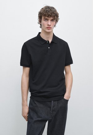 Jonge man met krullend haar, gekleed in een zwart poloshirt met korte mouwen en donkere jeans, staand met één hand in de zak tegen een effen achtergrond.