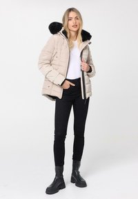 Chaqueta de plumas beige con capucha forrada de piel negra, con diseño acolchado y cremalleras, combinada con pantalones negros y botas negras gruesas.