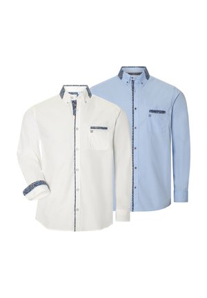 Mann trägt hellblaues Hemd mit Knöpfen und gemustertem Besatz sowie beige Hose, schaut seitlich mit Händen in den Taschen.
