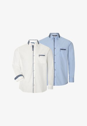 Mann trägt hellblaues Hemd mit Knöpfen und gemustertem Besatz sowie beige Hose, schaut seitlich mit Händen in den Taschen.