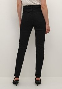 Jeans skinny neri a vita alta con una finitura in denim testurizzato, due tasche sul retro e una cintura liscia, abbinati a tacchi a stiletto neri.