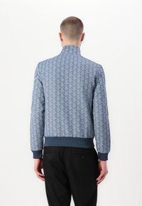 Jachetă bomber cu model albastru, având un design geometric, manșete și guler ribbed, cu o talie solidă de culoare albastru închis.