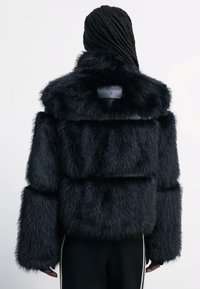 Veste cropped en fausse fourrure noire avec un large col, des manches volumineuses et des sections horizontales. Présente un détail logo au niveau du col.