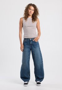 Striped sleeveless top in white and burgundy, paired with loose-fit blue denim jeans featuring wide legs and front pockets. Black sneakers.