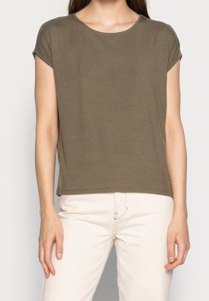 Kvinde iført en olivengrøn kortærmet t-shirt med rund hals kombineret med højtailede off-white bukser, stående foran en ensfarvet baggrund.