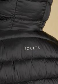 Detailný záber na čiernu prešívanú bundu zozadu s logom značky Joules vytlačeným bielymi písmenami.