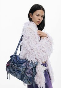 Mladá žena s dlhými čiernymi vlasmi, ktorá má na sebe textúrovaný levanduľový sveter, fialové nohavice a nesie veľkú modrú vzorovanú kabelku cez rameno.