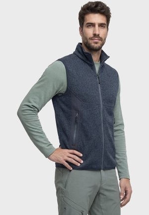 Mann trägt eine dunkelgraue Fleeceweste mit Reißverschluss über einem hellgrünen Langarmshirt und passenden grünen Hosen, posiert mit einer Hand in der Hüfte.