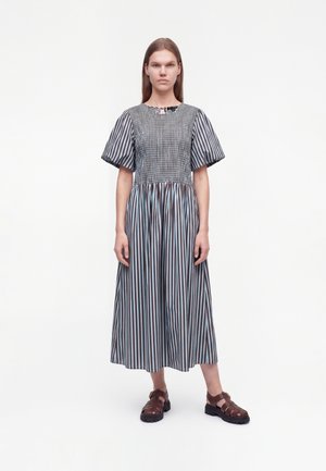 Mujer de pie con un vestido midi de rayas con mangas abullonadas y sandalias marrones gruesas con hebilla, sobre un fondo blanco liso.