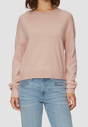 Femme portant un pull léger à manches longues rose pâle et un jean taille haute bleu clair à coupe décontractée, debout devant un fond uni.