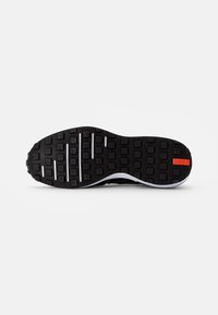 Semelle de chaussure noire avec motif quadrillé, bord de semelle intermédiaire blanc et petit détail rectangulaire rouge près du talon sur un fond clair.