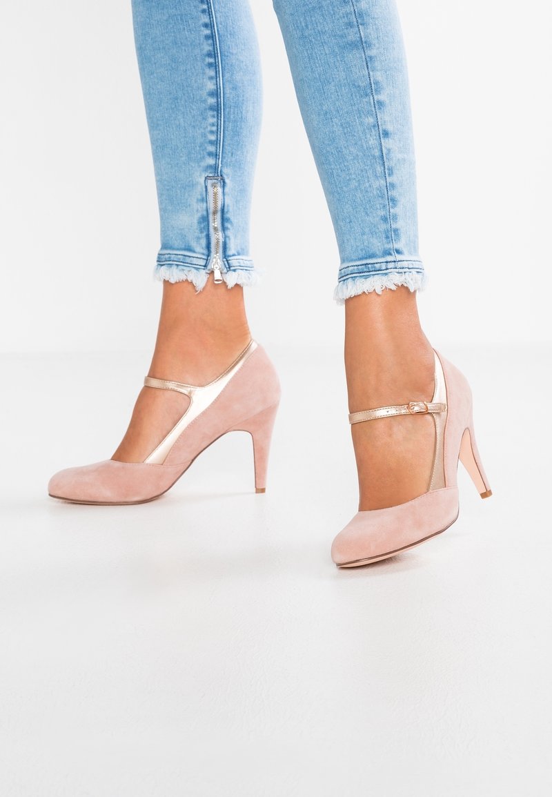 Růžové semišové lodičky na vysokém podpatku s designem T-strap, které mají kovový akcent. Kombinované se světle modrými skinny džíny s roztrhaným lemem.