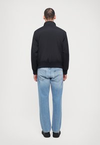Sort polstret jakke med høj krave, ribbede manchetter og kant, parret med lys blå straight-leg jeans og sorte sko. Bagfra.