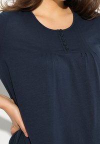 Navyblaues Hemd mit rundem Ausschnitt, senkrechten Falten und einer Knopfleiste mit fünf runden schwarzen Knöpfen. Glatte Stoffoberfläche.