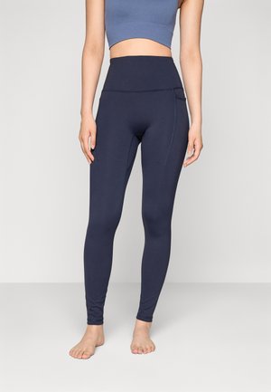 Leggings taille haute bleu marine fabriqués dans un matériau extensible avec une texture lisse, doté de poches latérales et de coutures plates pour le confort.