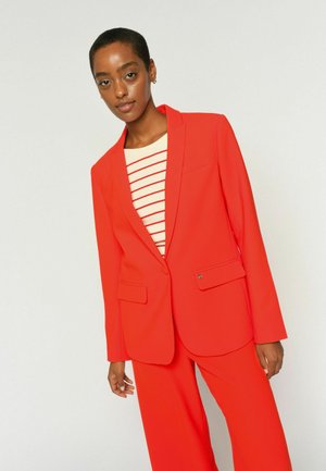 Mujer vistiendo un traje rojo brillante con un blazer de un solo botón y pantalones de pierna ancha sobre una camiseta blanca con rayas horizontales rojas.