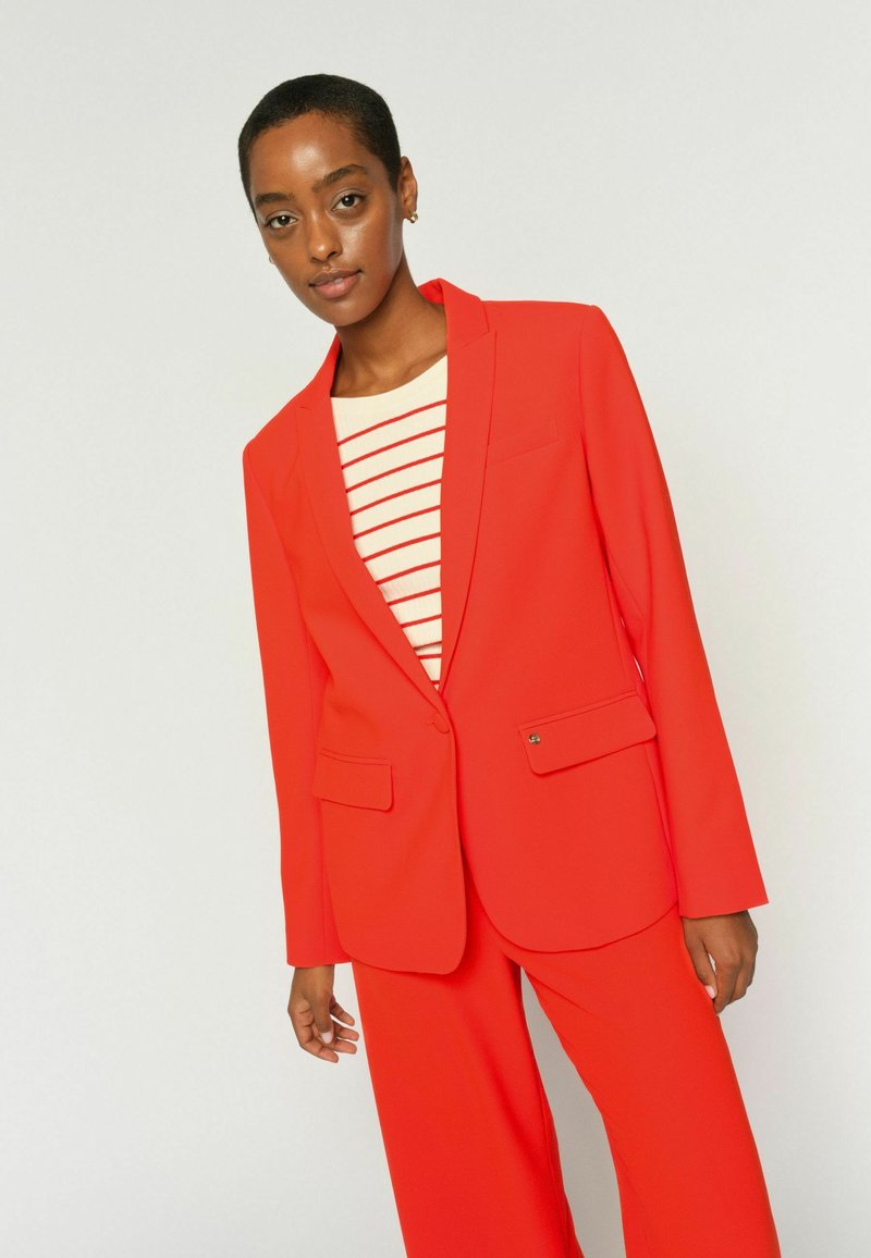 Žena v jasne červenom obleku s jedno-gombíkovým sako a nohavicami širokých nohavíc cez biely top s červenými horizontálnymi pruhmi.