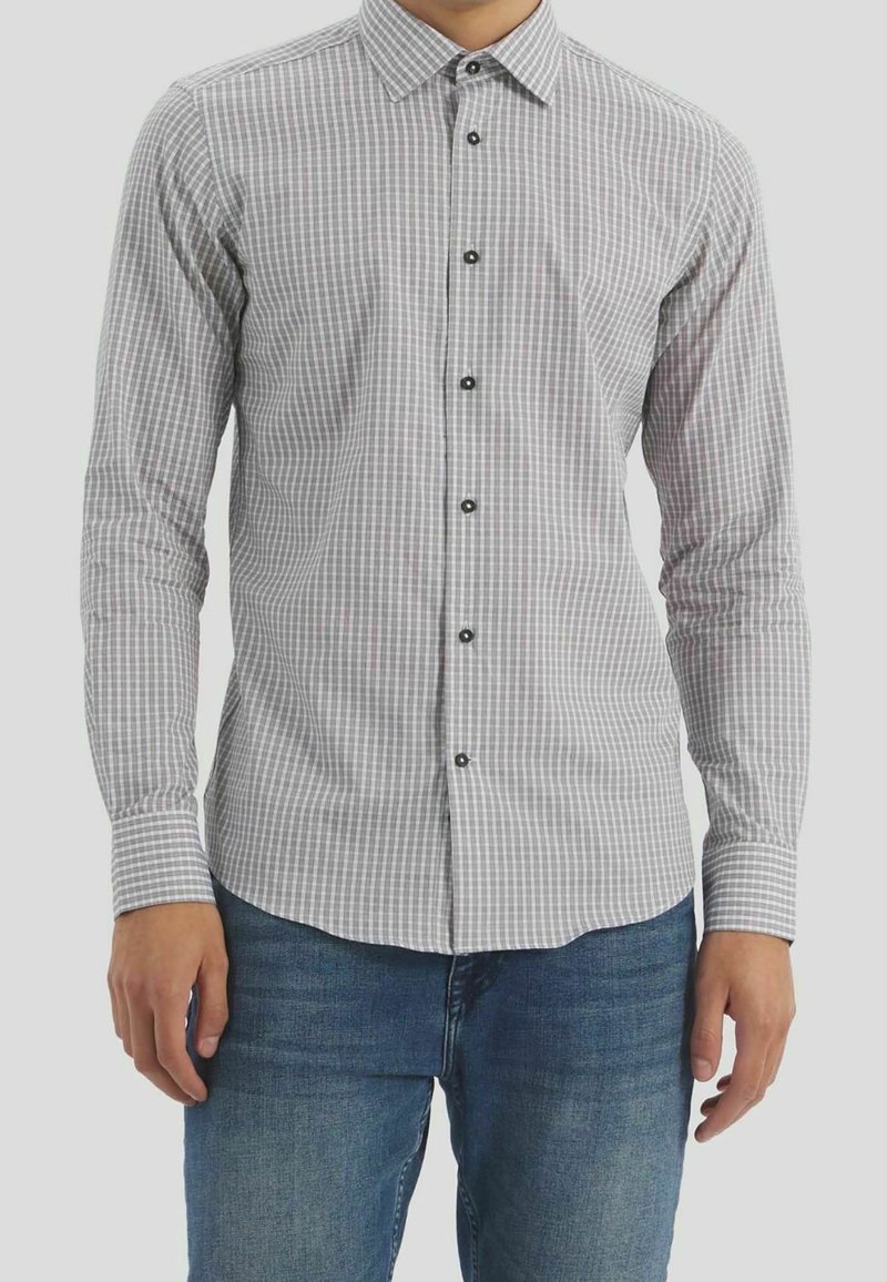 Chemise à carreaux gris et blanc avec col à boutons, manches longues et boutons noirs. Portée avec un jean bleu, présentant un ourlet arrondi.