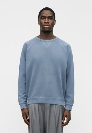 Mężczyzna ubrany w stonowaną niebieską bluzę crewneck i szare spodnie, stojący z rozluźnionymi ramionami na jednolitym białym tle.