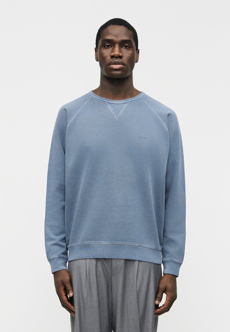 Homme portant un sweat-shirt à col rond bleu pâle et un pantalon gris, debout avec les bras détendus sur un fond blanc uni.