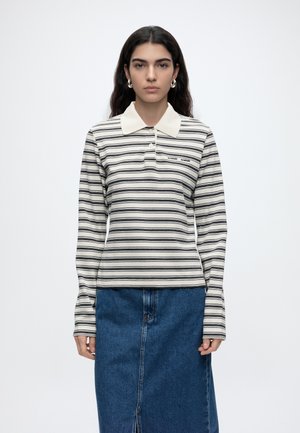 Frau mit dunklen Haaren, die silberne Creolen, ein weißes und schwarz gestreiftes Langarm-Poloshirt und einen blauen Jeansrock trägt.