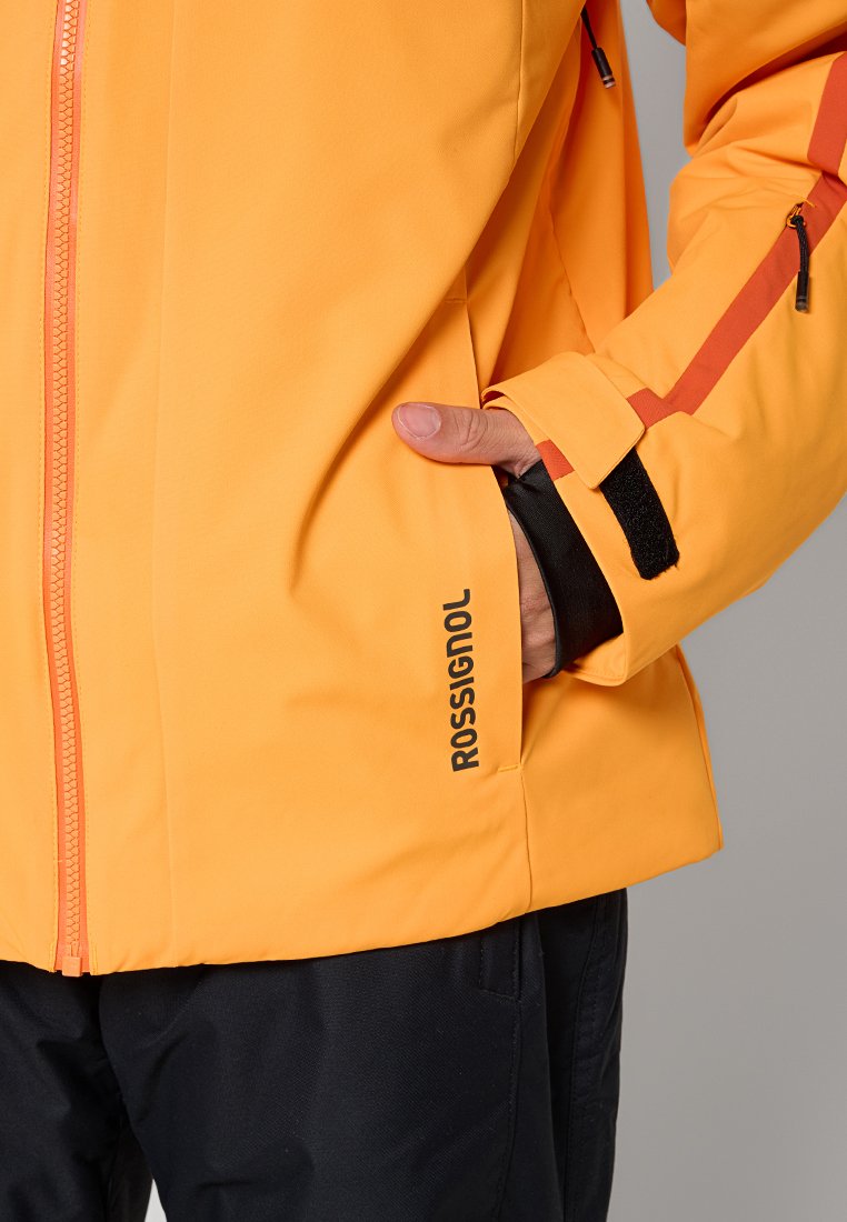 Jasnopomarańczowa wodoodporna kurtka z zamkiem z przodu, czarnymi akcentami i logotypem. Ma kieszeń z boku oraz regulowane mankiety.